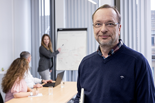 Das Bild zeigt einen Mann mit Schreibblock, die lächelnd in die Kamera schaut. Im Hintergrund sind drei Mitarbeitende verschwommen im Gespräch zu sehen.